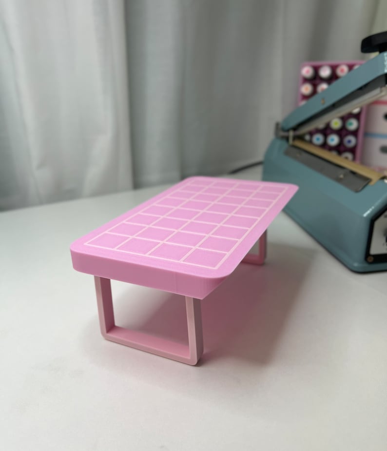May include: A pink table with a grid pattern on the top and a white border. The table has a simple design with two legs on each side.