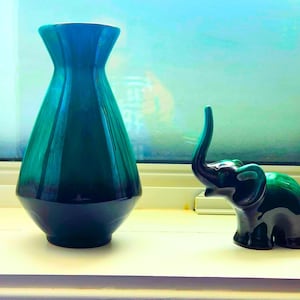 May include: A teal ceramic vase and a small green elephant ornament are displayed. The vase has a flared top and a dark base. The elephant has its trunk raised. The objects are on a white surface.