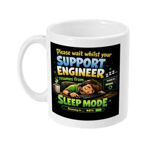 Może przedstawiać: Biały ceramiczny kubek z humorystyczną grafiką. Projekt przedstawia śpiącą osobę przy komputerze z napisem "Please wait whilst your SUPPORT ENGINEER resumes from SLEEP MODE". Kubek ma zakrzywiony uchwyt.