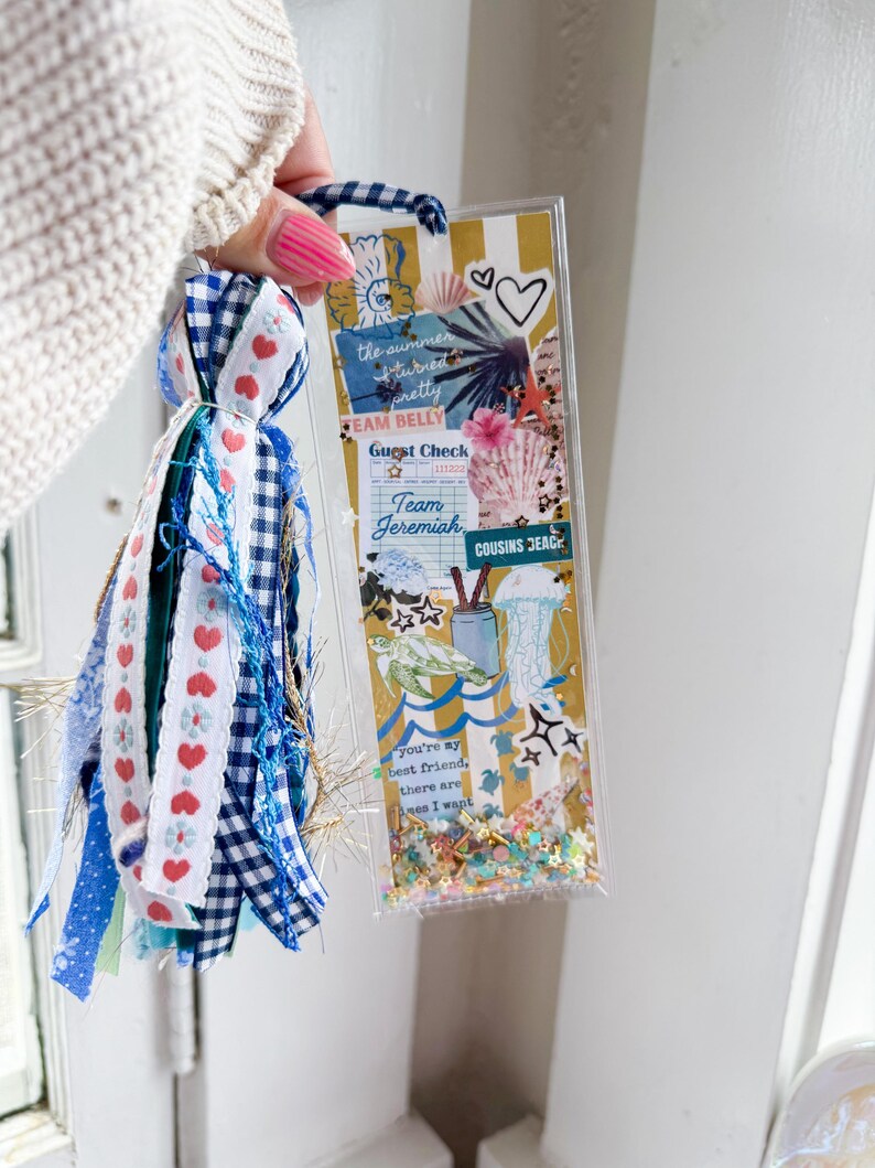 May include: A clear, rectangular bookmark with a tassel. The bookmark features a collage of beach-themed images and text, including "the summer turned pretty" and "Team Jeremiah." The tassel is made of blue, white, and red ribbons.