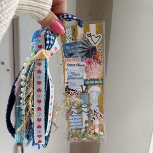 May include: A handmade bookmark with a clear plastic front filled with decorative elements. The bookmark features a blue and white gingham ribbon loop and a tassel with ribbons, beads, and a seashell. Text includes "the summer turned pretty" and "Team Belly".