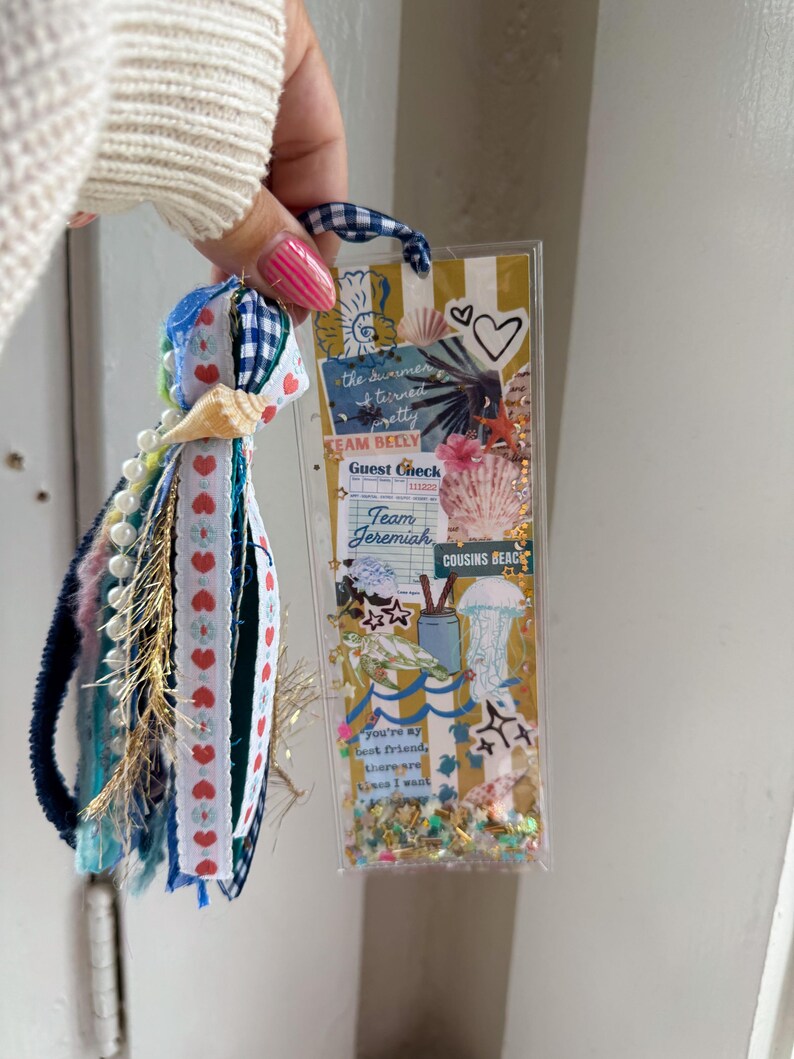 May include: A handmade bookmark with a clear plastic front filled with decorative elements. The bookmark features a blue and white gingham ribbon loop and a tassel with ribbons, beads, and a seashell. Text includes "the summer turned pretty" and "Team Belly".
