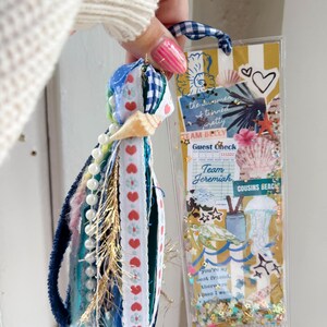 May include: A handmade bookmark with a tassel and a clear plastic sleeve. The tassel features blue, white, and red ribbons, beads, and a seashell. The bookmark has a striped background with text that reads "Team Jeremiah" and "Cousins Beach."