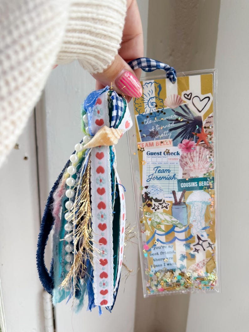 May include: A handmade bookmark with a tassel and a clear plastic sleeve. The tassel features blue, white, and red ribbons, beads, and a seashell. The bookmark has a striped background with text that reads "Team Jeremiah" and "Cousins Beach."