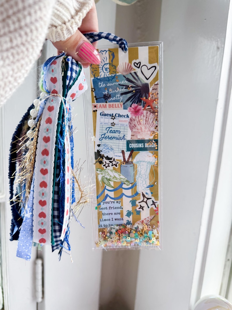 May include: A decorative bookmark with a tassel. The bookmark features a collage of beach-themed images, including seashells, a turtle, and the text "the summer turned pretty". The tassel includes blue, white, and red ribbons.