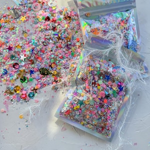 May include: Assortment of colorful glitter and sequins in clear plastic bags. The glitter and sequins are in various shapes, including stars, hearts, and circles. The bags are sealed with a zipper.