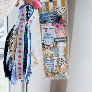 May include: A clear, rectangular bookmark with a beach theme, featuring seashells, a turtle, and the text "the summer turned pretty". It has a tassel with blue, white, and red ribbons and beads. The bookmark is held by a person.