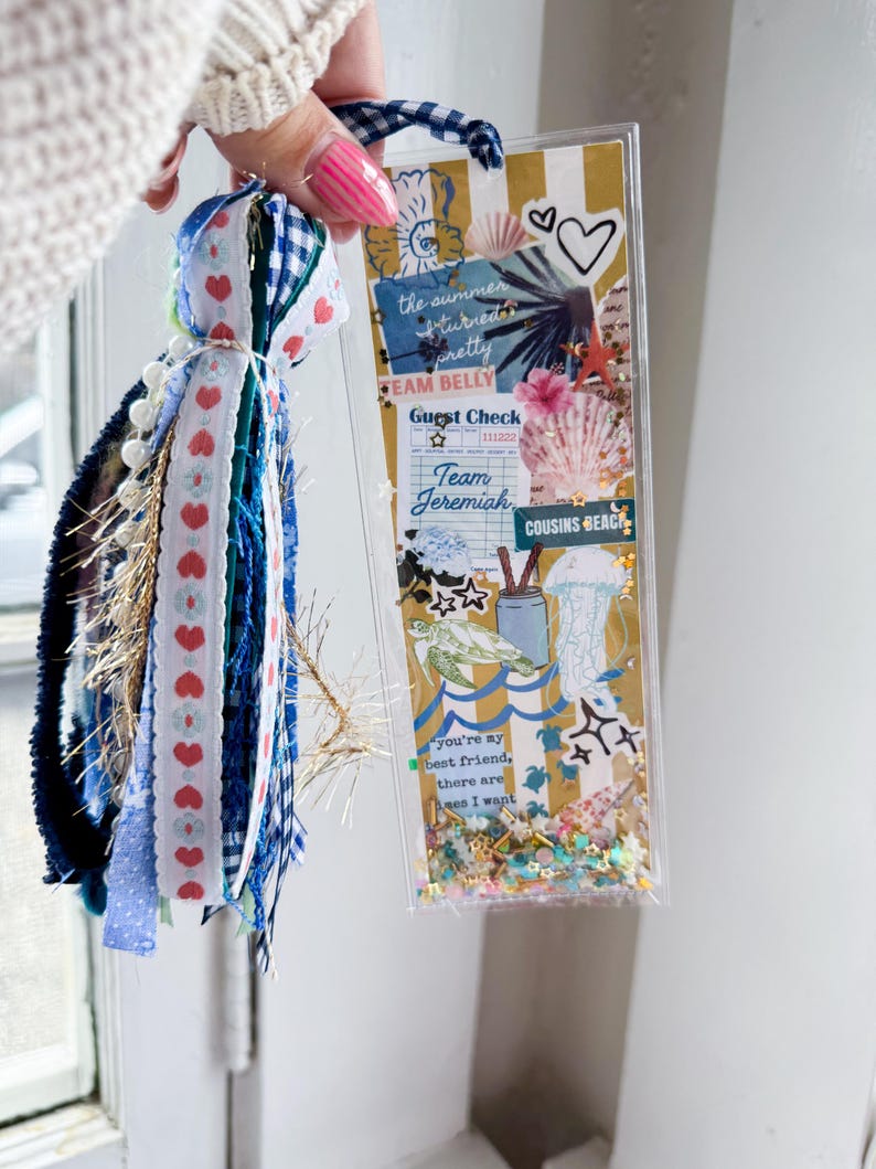 May include: A clear, rectangular bookmark with a beach theme, featuring seashells, a turtle, and the text "the summer turned pretty". It has a tassel with blue, white, and red ribbons and beads. The bookmark is held by a person.