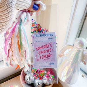 May include: A clear acrylic gift box filled with colorful confetti, disco balls, and a "Teacher Check" note that reads "Someone's Favorite Teacher." A decorative tassel with ribbons is held above the box.