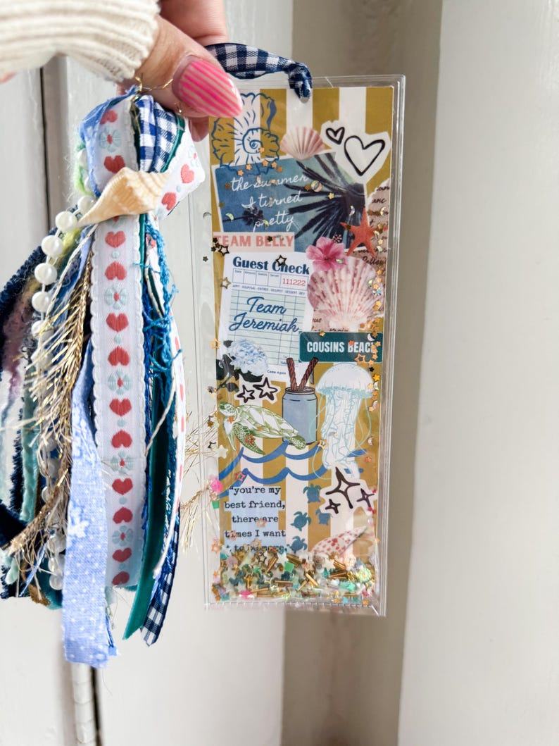 May include: A clear, rectangular shaker bookmark with a beach theme, featuring seashells, hearts, and the text "the summer & turned pretty". The bookmark has a blue and white gingham ribbon and a tassel with ribbons and beads.