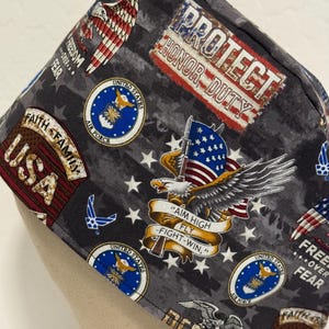 May include: A gray and black fabric scrub hat with a patriotic theme. It features a variety of images including the American flag, eagles, stars, and the words "Protect Honor Duty" and "Faith Family USA".