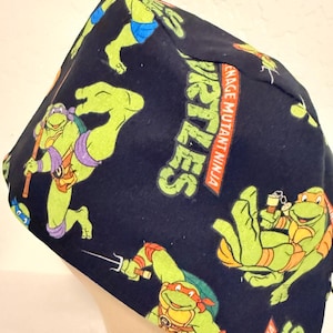 May include: A navy blue surgical scrub cap featuring a repeating pattern of the Teenage Mutant Ninja Turtles. The design includes the turtles in various action poses, with the words "Teenage Mutant Ninja Turtles" printed on the side.