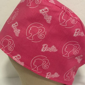 May include: A pink surgical scrub cap featuring a repeating pattern of the word "Barbie" and a silhouette of Barbie's profile in white. The cap has a tie closure and is made of a soft, lightweight fabric.