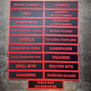 May include: A collection of black and red labels, each with a different tool category name in red capital letters. Categories include "Dado Blades", "Clamps", "Jigs", "Router Accessories", "Sandpaper", and more. The labels are arranged in a grid.