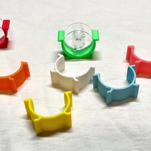 May include: A collection of colorful plastic clips with clear, circular inserts. The clips are in various colors, including red, green, orange, white, blue, yellow, and tan. The objects are arranged on a white surface.