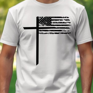 Puede incluir: Camiseta blanca con un diseño gráfico negro. El diseño combina una cruz con una bandera estadounidense desgastada. Las estrellas y las rayas de la bandera son visibles, creando un símbolo patriótico y religioso. La camiseta es una prenda casual.