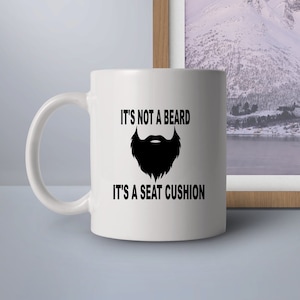 Puede incluir: Taza de cerámica blanca con un gráfico negro de una barba y el texto "No es una barba, es un cojín de asiento."
