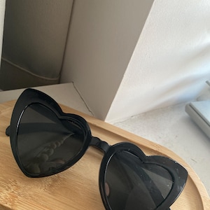 May include: Black heart-shaped sunglasses with dark lenses. The sunglasses are resting on a light brown wooden tray. The frames are thick and the lenses are dark, providing a stylish and protective accessory.