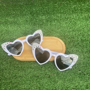 May include: Three pairs of white heart-shaped sunglasses with black lenses are displayed on a wooden tray. Each pair has text on the frame: "PROM", "LILAH", and "FREYA" in silver letters. The sunglasses are set against a green background.