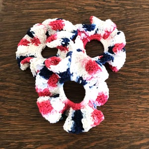 May include: Three handmade crochet scrunchies in a patriotic color scheme of red, white, and blue. The scrunchies are circular with a hole in the center and a ruffled edge. The scrunchies are on a wooden surface.