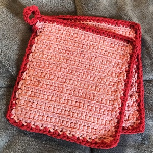 May include: Two handmade crochet potholders. The potholders are square, with a gradient of pink hues, and a solid red border. A loop is attached to one corner for hanging. The potholders are approximately 8 inches square.