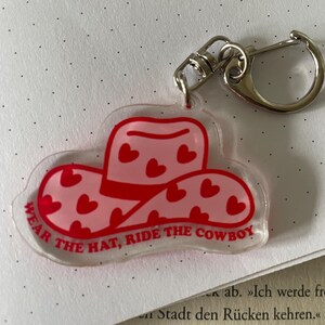Könnte beinhalten: Transparenter Acryl-Schlüsselanhänger in Form eines Cowboyhuts. Der Hut ist rot mit rosa Herzakzenten und dem Text "WEAR THE HAT, RIDE THE COWBOY". Er hat einen silberfarbenen Verschluss.
