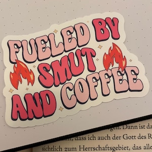 Könnte beinhalten: Rosa und roter Aufkleber mit dem Text "Fueled by Smut and Coffee" mit Flammen auf beiden Seiten des Textes.