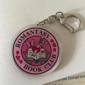 Könnte beinhalten: Ein klarer, runder Schlüsselanhänger mit einem rosa Aufdruck "Romantasy Book Club". Das Design zeigt ein Schwert durch ein Herz, ein offenes Buch, Schmetterlinge und Herzen. Er hat einen silbernen Verschluss.