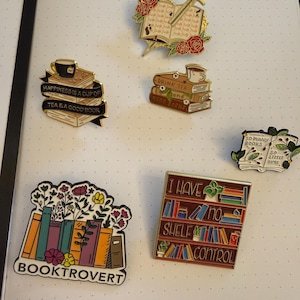 Könnte beinhalten: Eine Sammlung von Emaille-Anstecknadeln mit Buch- und Teemotiven. Designs umfassen Bücherstapel mit Teetassen, offene Bücher mit Schwertern und ein Bücherregal mit dem Text "I have no shelf control".