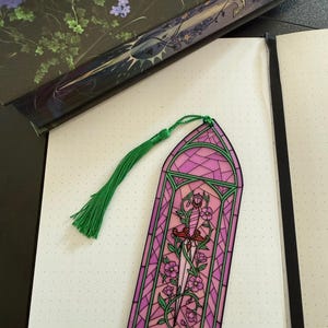 May include: A rectangular bookmark featuring a stained glass design in shades of pink and green. The artwork depicts a central dagger entwined with flowering vines, framed by a geometric border. A green silk tassel is attached to the top.