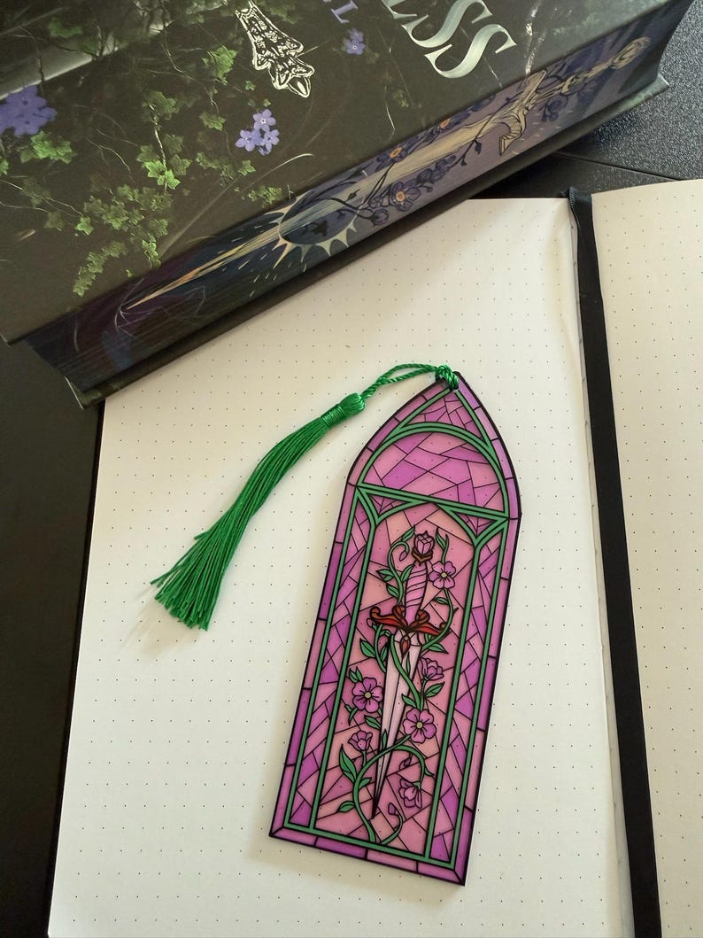May include: A rectangular bookmark featuring a stained glass design in shades of pink and green. The artwork depicts a central dagger entwined with flowering vines, framed by a geometric border. A green silk tassel is attached to the top.