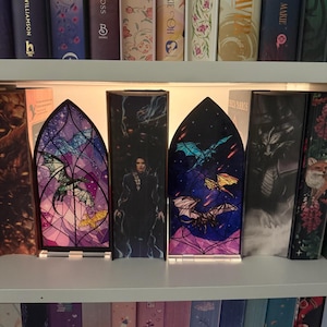 Könnte beinhalten: Zwei bogenförmige Buchstützen im Buntglas-Stil mit Drachenillustrationen. Das linke Stück zeigt einen grünen und blauen Drachen vor violettem Hintergrund. Das rechte Stück zeigt blaue und gelbe Drachen vor einem dunkelblauen Sternenhimmel.