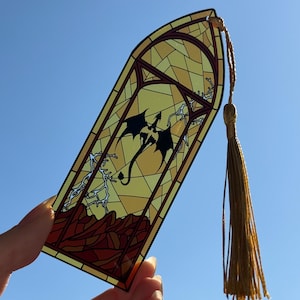 May include: A translucent bookmark featuring a stained glass design with a black dragon silhouette, lightning bolts, and a red mountain landscape. The bookmark has a pointed top and includes a long, gold-colored decorative tassel attached at the top.