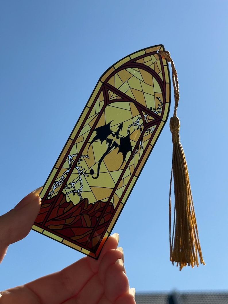 May include: A translucent bookmark featuring a stained glass design with a black dragon silhouette, lightning bolts, and a red mountain landscape. The bookmark has a pointed top and includes a long, gold-colored decorative tassel attached at the top.