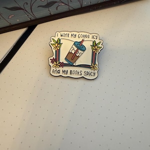 Könnte beinhalten: Ein quadratischer Emaille-Pin mit abgerundeten Kanten, der ein offenes Buch, einen Eiskaffee mit rotem Strohhalm und gelbe Blumen zeigt. Der Text lautet I WANT MY COFFEE ICY und AND MY BOOKS SPICY in schwarzer Schrift auf weißem Grund.