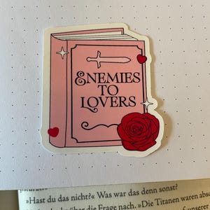 Könnte beinhalten: Ein rosa Buch-Aufkleber mit einem Schwert und einer roten Rose. Der Buchtitel lautet "Enemies to Lovers".