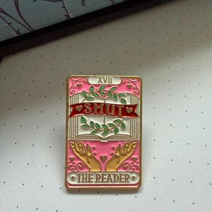 Könnte beinhalten: Ein rosafarbener und goldfarbener Emaille-Pin mit einem Tarotkarten-Design. Der Pin trägt die Aufschrift "SMUT" in der Mitte und "THE READER" unten. Das Design umfasst Hände, ein Buch und dekorative Elemente.