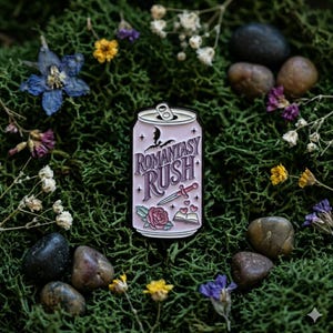 Könnte beinhalten: Ein rosa Emaille-Pin in Form einer Getränkedose mit dem Text "Romantasy Rush" und Illustrationen einer Fledermaus, eines Schwertes, einer Rose und eines Buches. Der Pin ist auf grünem Moos platziert, umgeben von Blumen und kleinen Steinen.