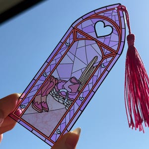 May include: A translucent purple bookmark shaped like a gothic arch window with a stained glass pattern. The design features a pair of pink cowboy boots, a hat, and a horseshoe. A bright pink tassel is attached to the top of the bookmark.