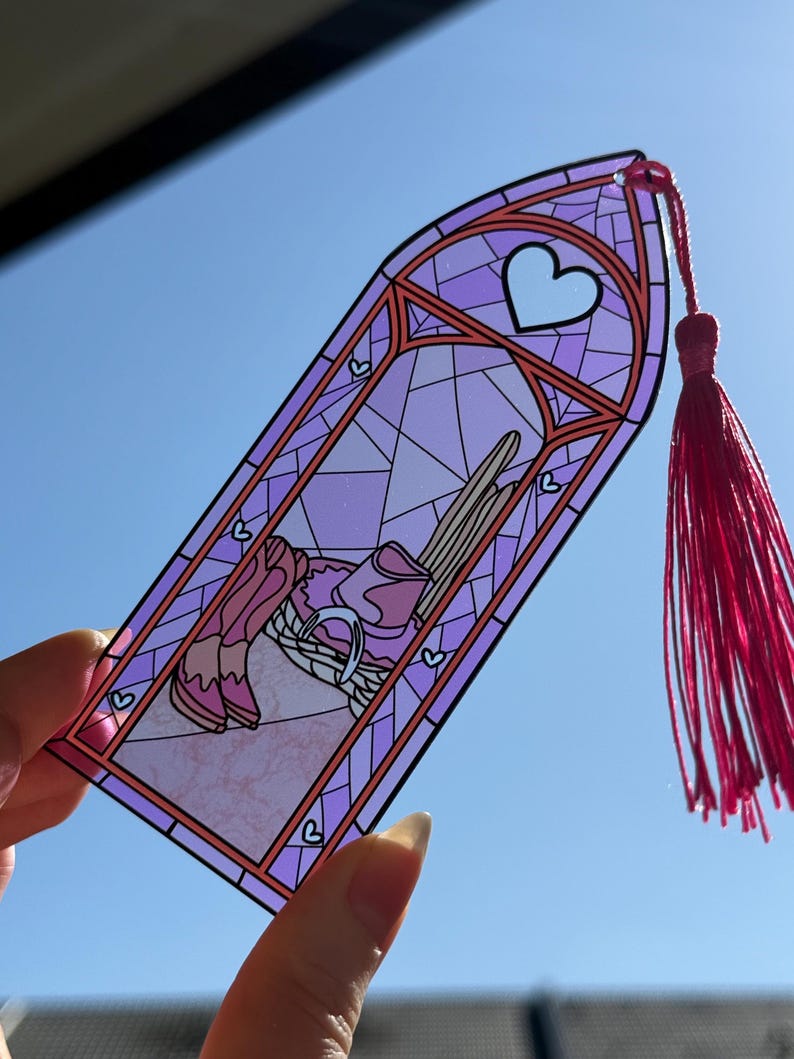 May include: A translucent purple bookmark shaped like a gothic arch window with a stained glass pattern. The design features a pair of pink cowboy boots, a hat, and a horseshoe. A bright pink tassel is attached to the top of the bookmark.