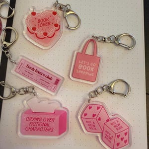 Könnte beinhalten: Fünf durchsichtige Acryl-Schlüsselanhänger mit silberfarbenen Metallringen. Die Motive umfassen eine herzförmige rosa Torte mit der Aufschrift BOOK LOVER, eine rosa Tragetasche mit LET'S GO BOOK SHOPPING, ein Book lovers club Ticket, eine Taschentuchbox und zwei rosa Würfel mit Herzen.
