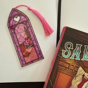 May include: A rectangular bookmark with a stained glass window design in shades of pink and purple, featuring a heart at the top and a western scene with boots and a hat at the bottom. The bookmark includes a pink tassel attached to the top.