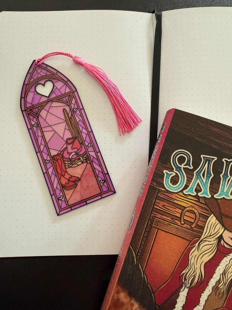 May include: A rectangular bookmark with a stained glass window design in shades of pink and purple, featuring a heart at the top and a western scene with boots and a hat at the bottom. The bookmark includes a pink tassel attached to the top.