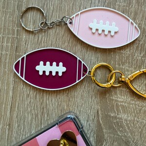 Könnte beinhalten: Zwei fußballförmige Schlüsselanhänger. Einer ist rosa mit weißen Akzenten, der andere weinrot mit weißen Akzenten. Beide haben ein weißes Fußballstichdesign. Die Schlüsselanhänger sind an silbernen und goldenen Schlüsselringen befestigt.