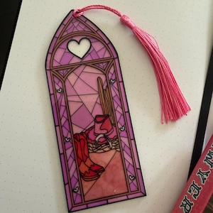 May include: A vertical bookmark featuring a stained-glass window design in shades of pink and purple. The illustration depicts a cowboy hat, a horseshoe, and a pair of boots. A white heart sits at the top, and a pink tassel is attached to the top edge.