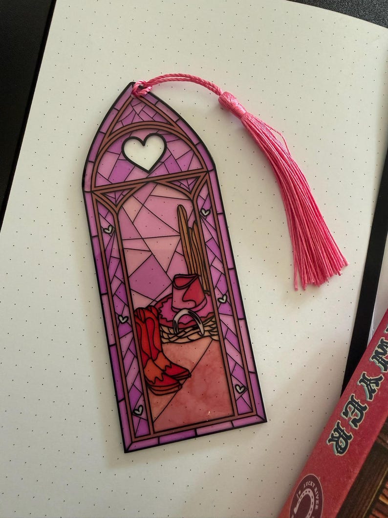 May include: A vertical bookmark featuring a stained-glass window design in shades of pink and purple. The illustration depicts a cowboy hat, a horseshoe, and a pair of boots. A white heart sits at the top, and a pink tassel is attached to the top edge.