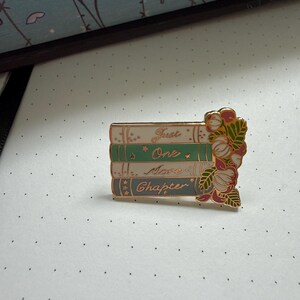 Pode incluir: Um broche de esmalte retangular com uma pilha de quatro livros nas cores branco, verde, azul-claro e azul-escuro. Os livros trazem a inscri&ccedil;&atilde;o dourada Just One More Chapter. Um grupo de flores cor-de-rosa e folhas verdes adorna o lado direito.
