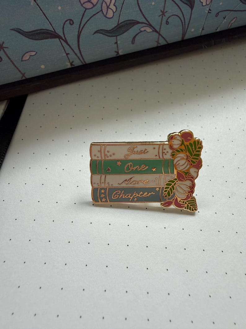 Pode incluir: Um broche de esmalte retangular com uma pilha de quatro livros nas cores branco, verde, azul-claro e azul-escuro. Os livros trazem a inscri&ccedil;&atilde;o dourada Just One More Chapter. Um grupo de flores cor-de-rosa e folhas verdes adorna o lado direito.