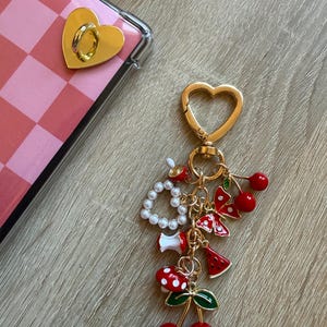 Puede incluir: Un llavero dorado en forma de corazón con varios dijes, que incluyen cerezas rojas, una seta, una rodaja de sandía y una cadena de perlas. El llavero está sujeto a un cierre dorado en forma de corazón. También se ve una funda de teléfono a cuadros rosa y blanco con un soporte para teléfono dorado en forma de corazón.