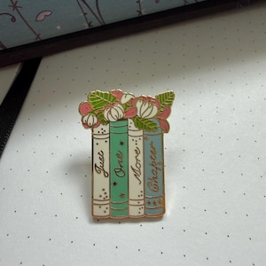 Pode incluir: Um broche de esmalte em tom dourado com quatro livros verticais com lombadas em tons pastel creme, verde menta e azul claro. Os livros s&atilde;o coroados com flores rosas e brancas e folhas verdes. O texto Just One More Chapter est&aacute; em dourado.